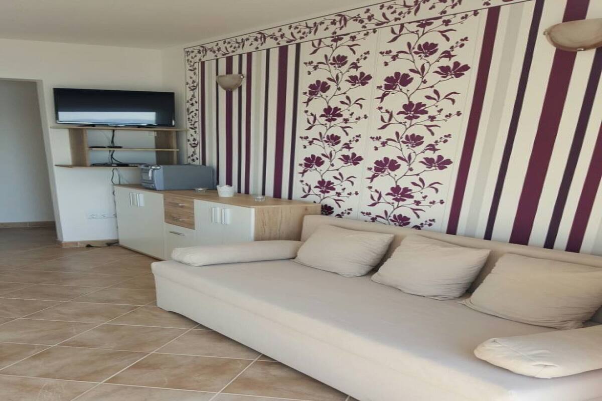 A living room with a beige sofa, a TV on a stand, a kitchen area, and a decorative wall with vertical stripes and floral patterns.