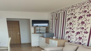 Un salon moderne avec un canapé beige, un téléviseur à écran plat fixé à un meuble en bois, et un coin repas avec une chaise blanche.