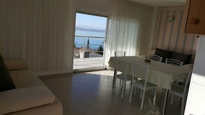 Uma sala de estar moderna com área de jantar, um sofá, uma mesa com cadeiras e vista para o mar através de uma porta de vidro.