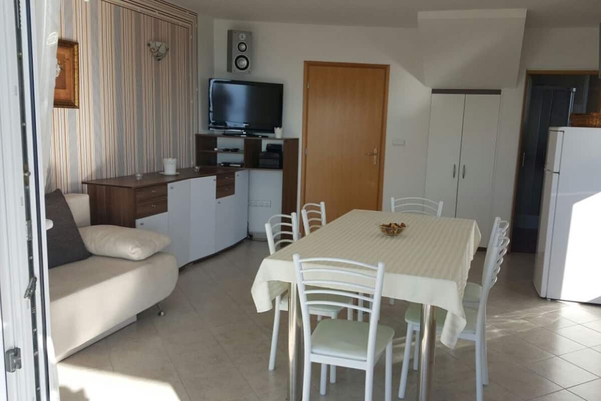 A compact kitchen with a dining table, chairs, a sofa, a television, and a refrigerator.