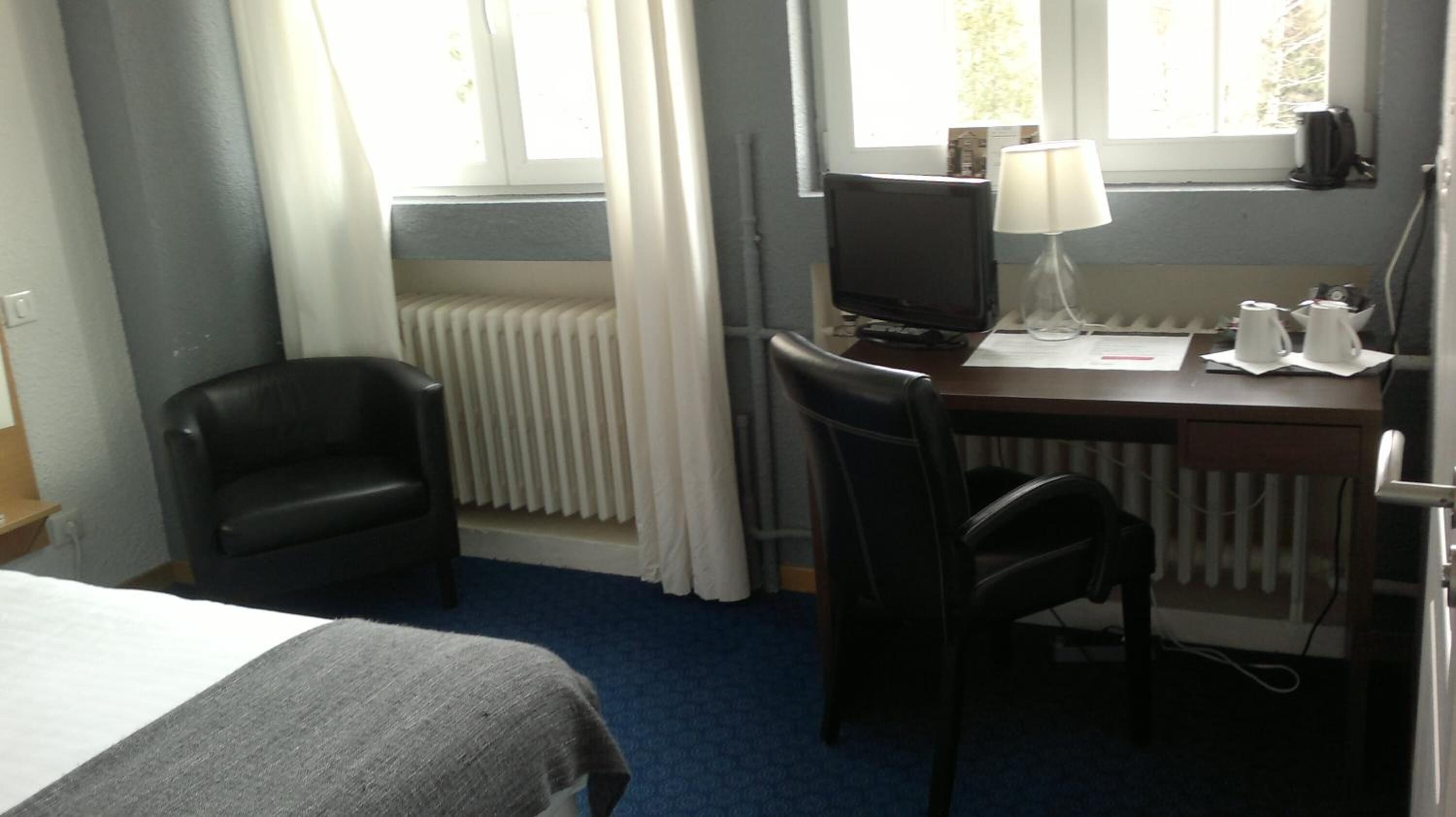 A hotel room with a bed, a desk with a computer, two chairs, a radiator, and a window with curtains.