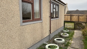 A house with a textured exterior, a window with a brown frame, and a pathway with tire planters. - UIST Travel Accommodation (South Uist)