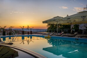 Zona de piscina amb gandules i para-sols, una piscina que reflecteix el capvespre i una vista del mar.