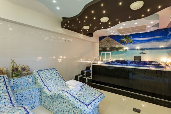 Un espace spa moderne doté d’un grand jacuzzi, d’un coin salon avec un sol en mosaïque et d’une fresque représentant une scène de plage tropicale.