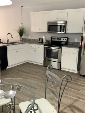 A modern kitchen with white cabinets, stainless steel appliances, a granite countertop, and a dining area with metal chairs. - The Executive Brook. (Greenville)