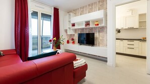 A modern living room with a red sofa, a TV mounted on a wooden wall, and a kitchen area visible through an open door. - GuestHost - Bright apartment included in a building of very recent construction (Milano)