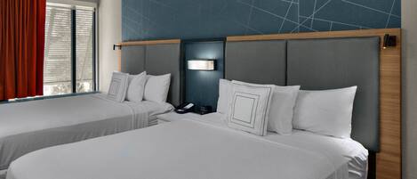 A hotel room with two beds, a large window with blinds, a wall with a geometric pattern, and a nightstand with a lamp.