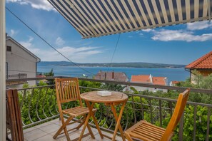 A balcony with two wooden chairs and a small table, overlooking a coastal view with houses and a clear sky.