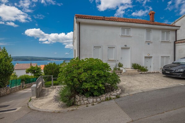 A two-story building with a white facade and red-tiled roof, a stone staircase leading to the entrance, and a view of the sea in the background.