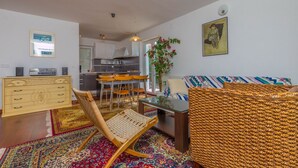 A living room with a sofa, a wicker chair, a dining table with chairs, a kitchen in the background, and a large window. - Apartment Maro (Crikvenica)