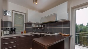 A modern kitchen with dark cabinetry, a central island, a built-in stove, and a window overlooking greenery. - Apartment Maro (Crikvenica)