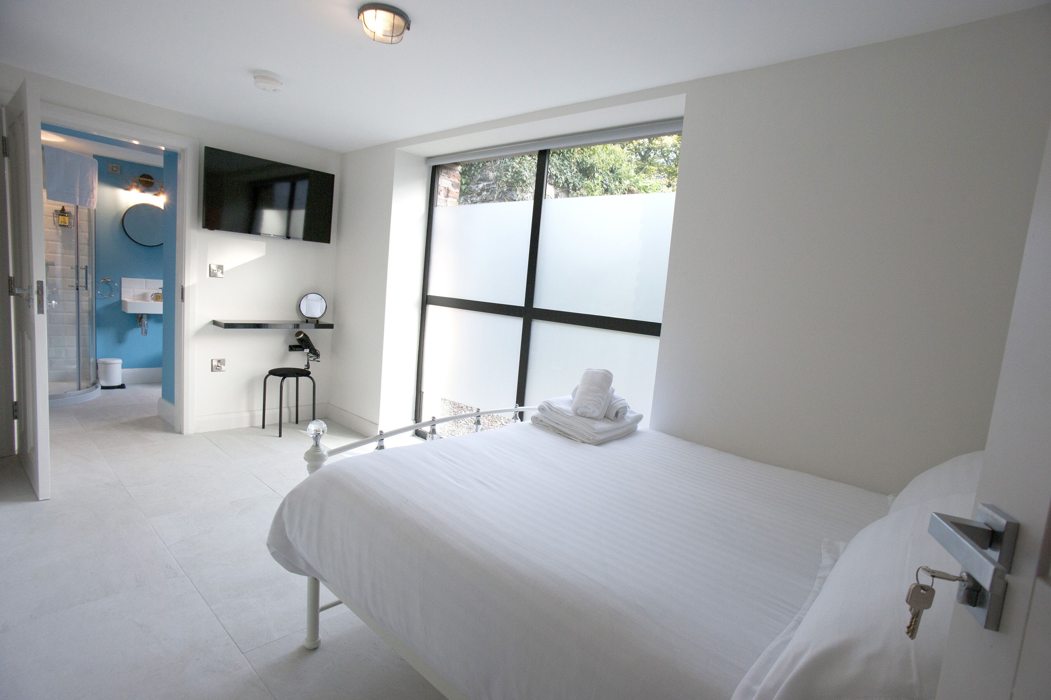 Une chambre d’hôtel moderne avec un grand lit, une salle de bain avec un mur d’accent bleu et une vue dégagée sur l’extérieur grâce à une grande fenêtre.