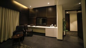 A modern hotel room with a dark wood panel wall, a flat-screen TV mounted above a white console table, a seating area with a small round table and two chairs, and a potted plant in the corner. - Hotel German Palace (Gandhinagar)