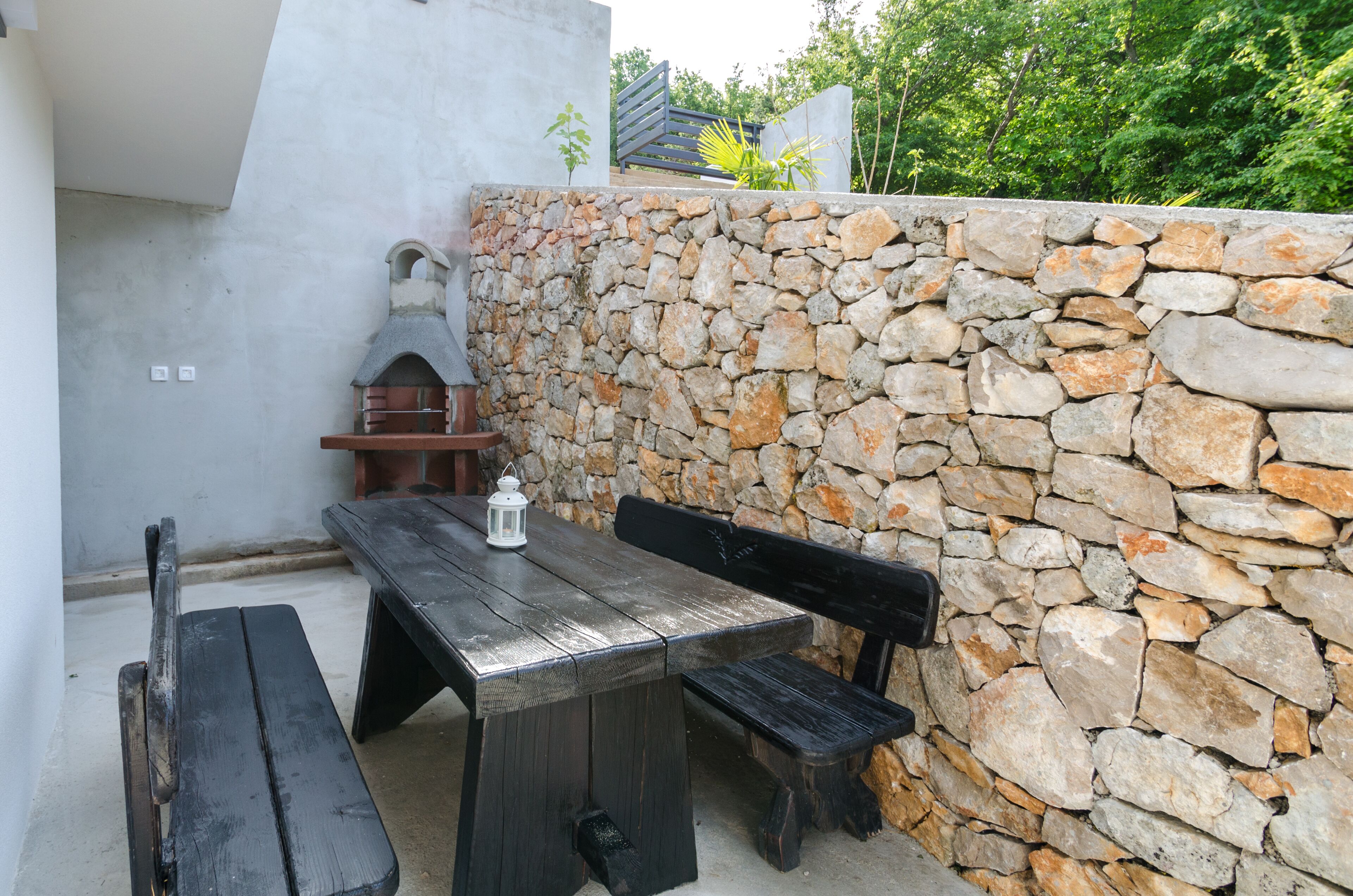 An outdoor patio with a stone wall, a wooden table, benches, and a brick oven.