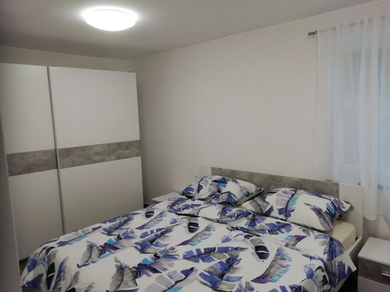 A bedroom with a bed, a nightstand, a wardrobe, and a window with curtains.