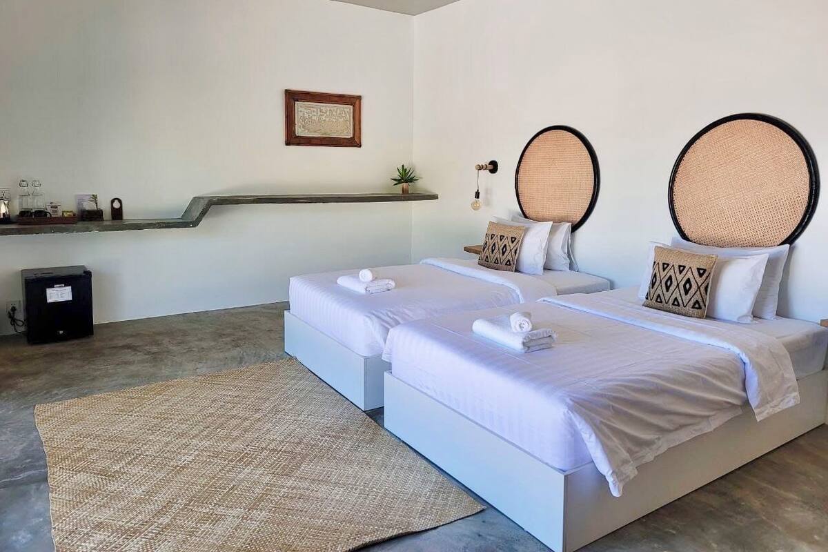 Habitación de hotel con dos camas, un escritorio pequeño, una estantería con objetos y un cuadro en la pared.