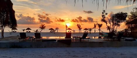 A beach scene with lounge chairs and umbrellas, a pool, and a sunset view.