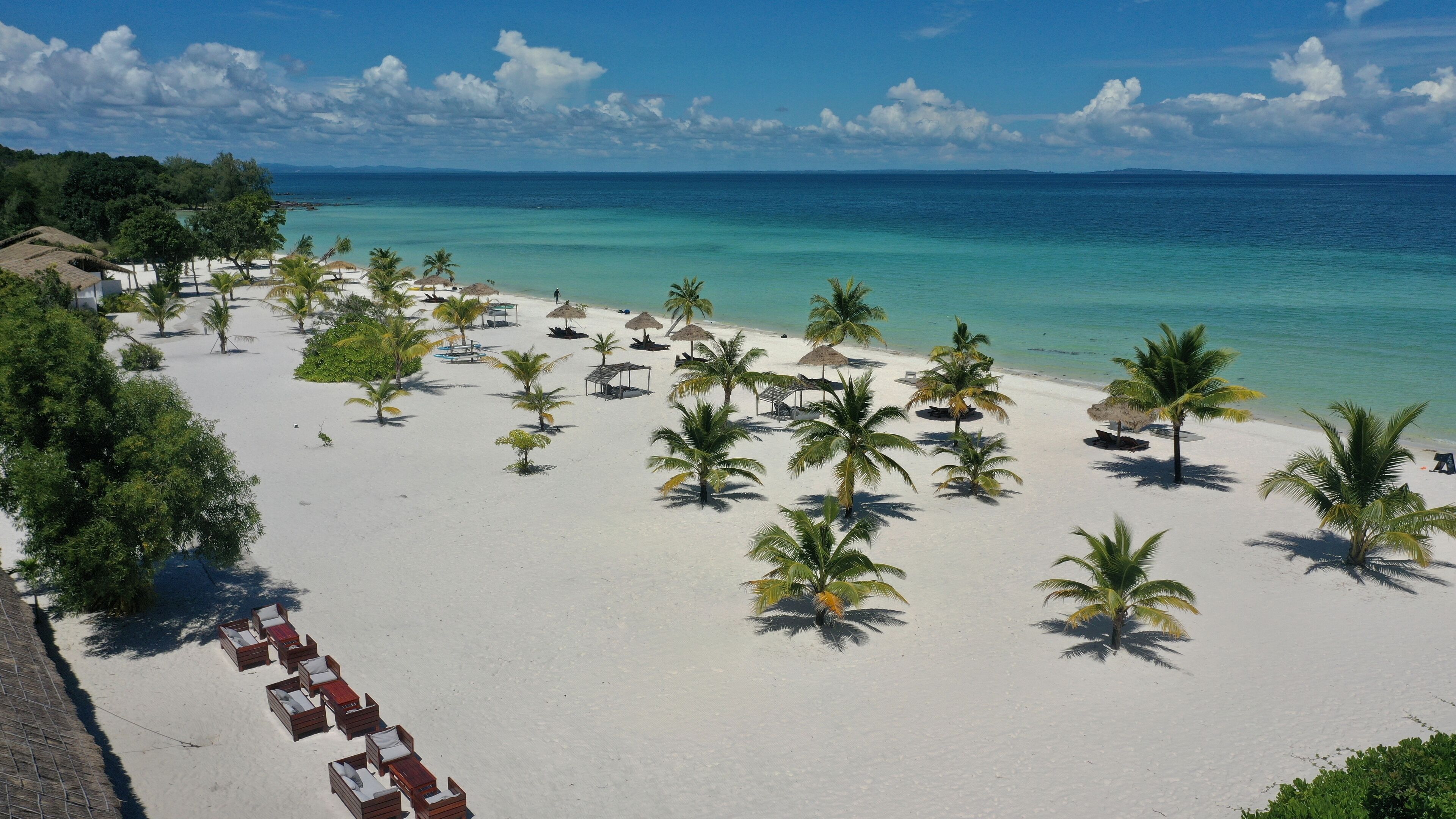 A beach with white sand, palm trees, and thatched umbrellas.
