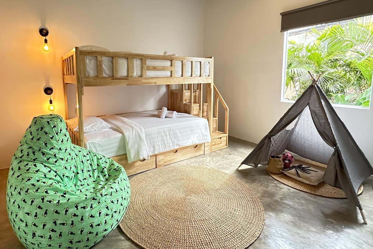 Dormitorio con literas, alfombra tejida, sillón de peluche verde, una tienda de campaña y una ventana con vistas a la vegetación.