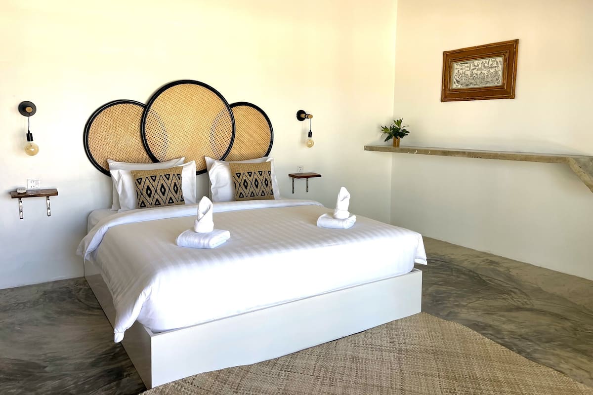 Una habitación de hotel moderna con una cama grande, cabecera de ratán y una pequeña estantería con una planta.