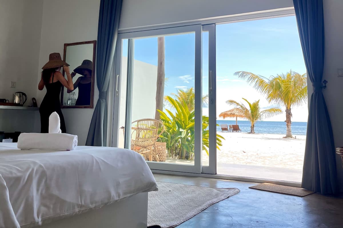 Habitación de hotel con cama, ventana grande con cortinas y vista a la playa, además de una silla afuera.