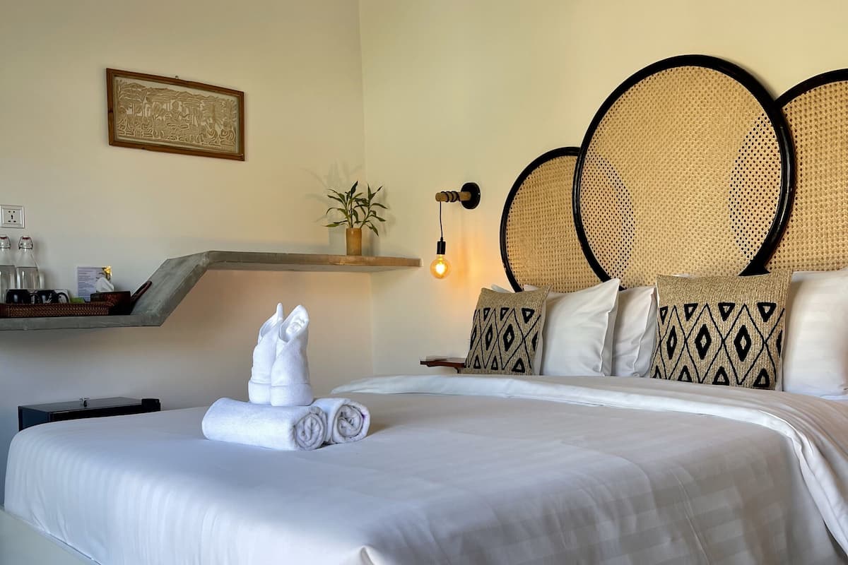 Una cama bien tendida con sábanas blancas y toallas enrolladas, una mesita de noche con una lámpara, un cuadro en la pared y una pequeña planta en maceta.