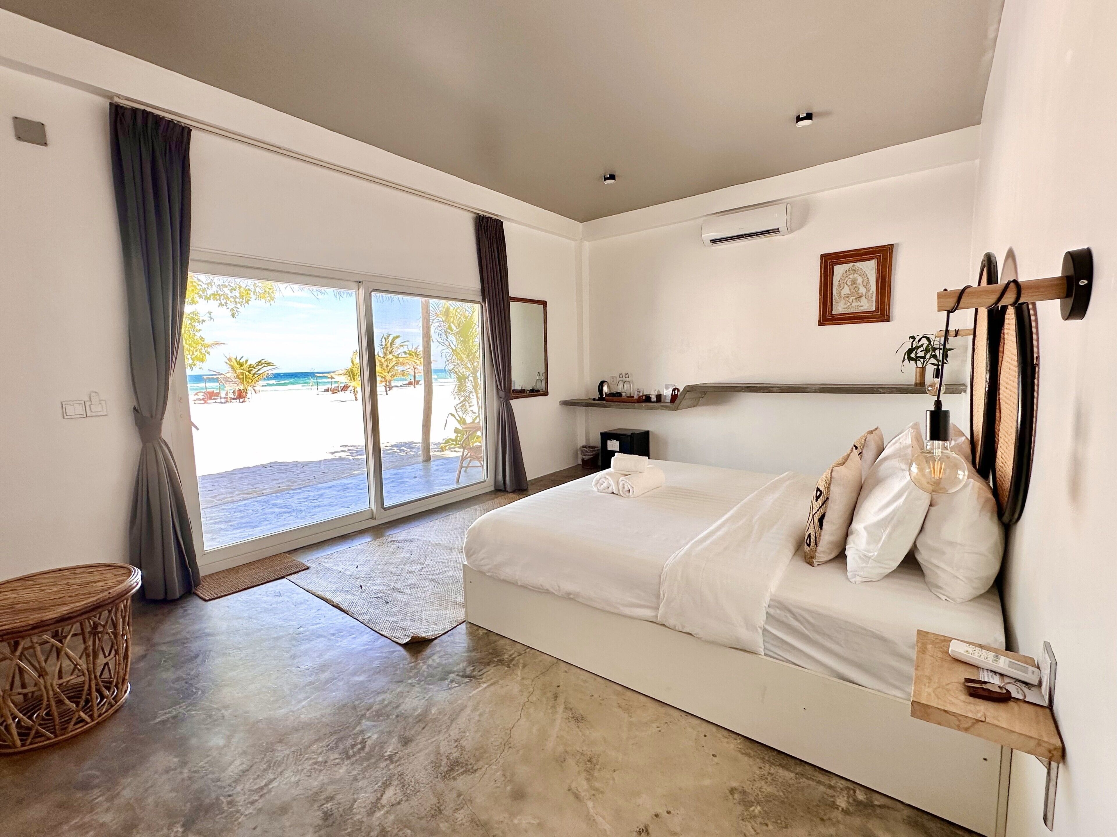 A bedroom with a large bed, a bedside table, a wall-mounted air conditioner, a shelf with items, and a view of the beach through a sliding door.