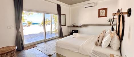 A bedroom with a large bed, a bedside table, a wall-mounted air conditioner, a shelf with items, and a view of the beach through a sliding door.