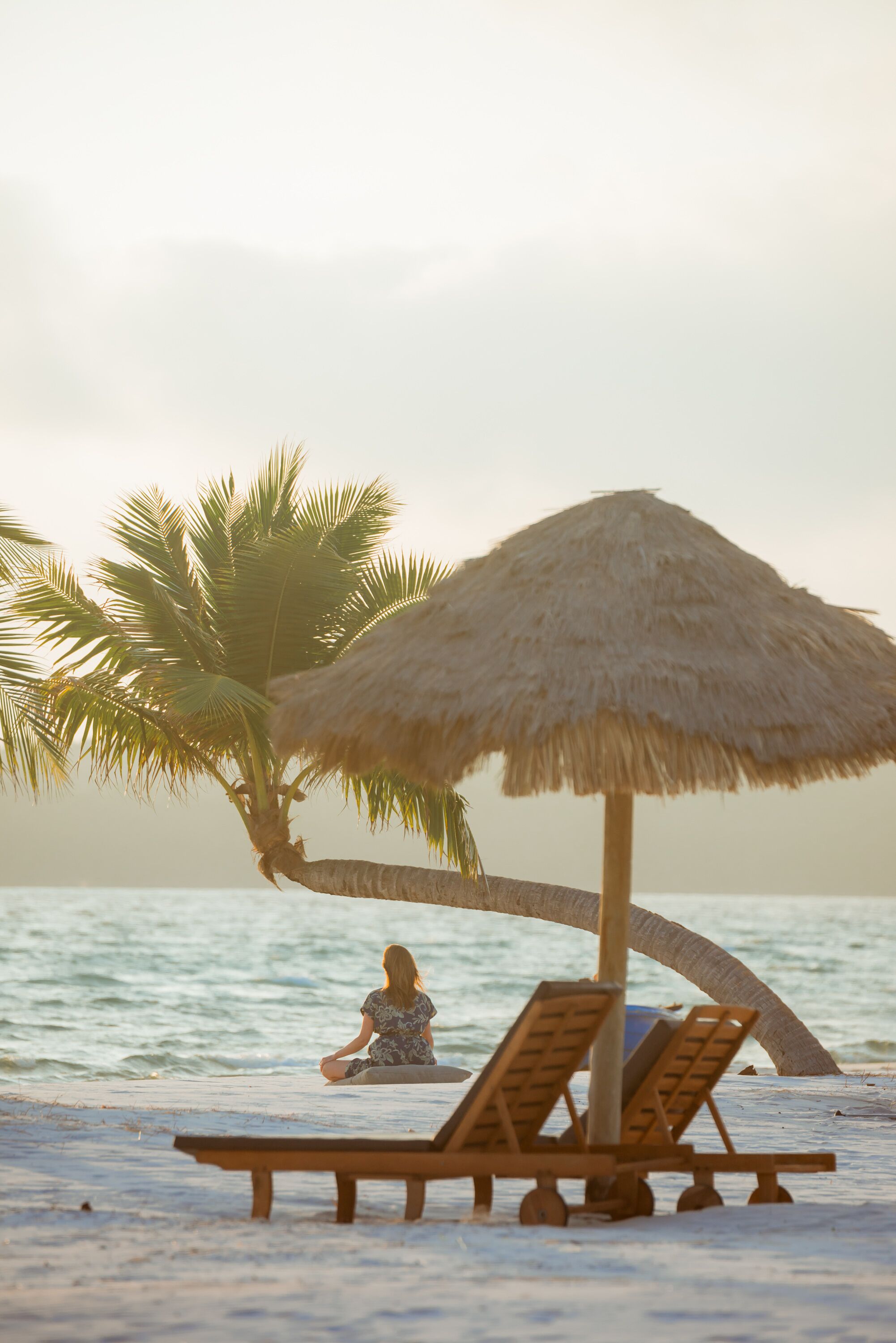 Escena de playa con una palmera, una sombrilla de paja y sillones de madera.