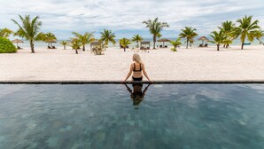 A woman standing at the edge of a pool with a beach and palm trees in the background.