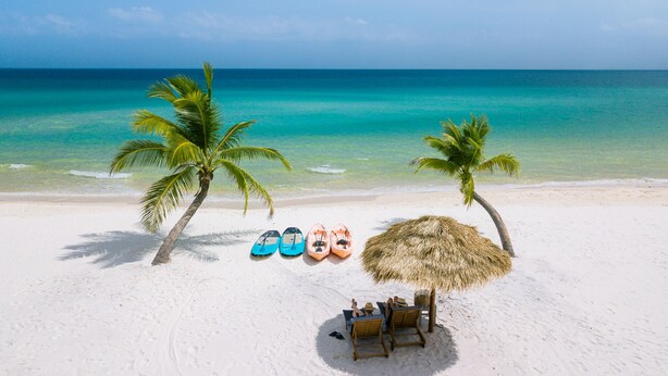 A beach with white sand, turquoise water, and palm trees. There are kayaks lined up and a thatched umbrella. - The Secret Garden Koh Rong (Koh Rong)