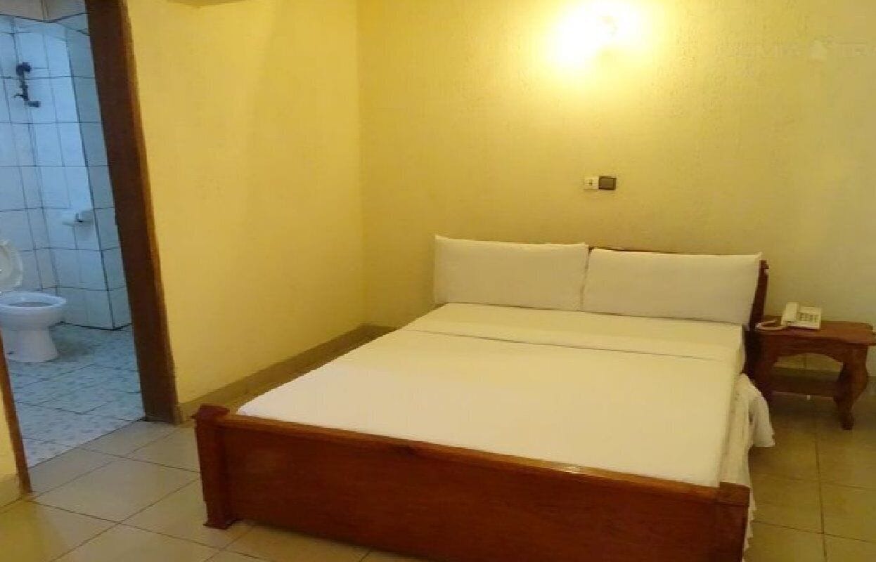 A small, single-bed room with a wooden frame, a white mattress, and a bedside table with a phone.