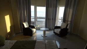 A hotel room with a bed, two armchairs, a small table, and a view of the sea through large windows. - FINI Downtown (Limbe)