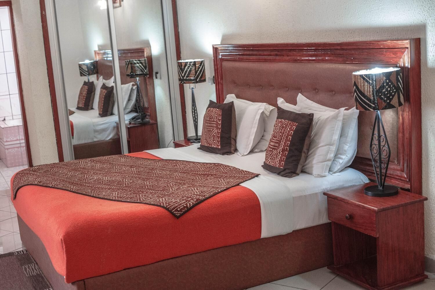 Cama bem arrumada com manta vermelha, várias almofadas e cabeceira de madeira. Há uma mesa de cabeceira com abajur e uma gaveta pequena.