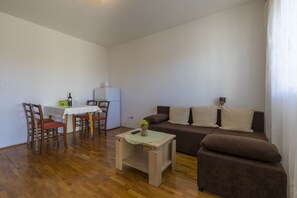A living room with a wooden floor, a brown sofa, a white table, and a dining area with chairs and a small refrigerator. - Apartments Milan (Crikvenica)