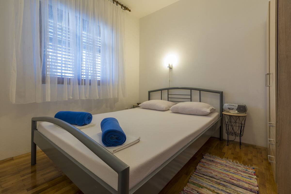 A single bed with a white mattress, two pillows, and a blue towel. There is a small bedside table with a radio. A window with white curtains allows natural light into the room.