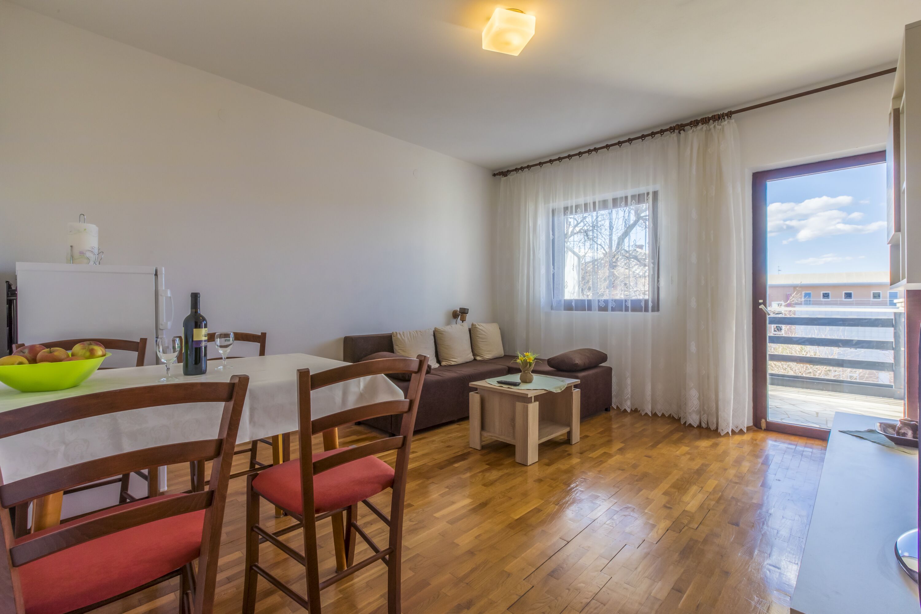 Un comedor con una mesa, sillas y un cuenco de fruta. Hay un sofá y una ventana con cortinas.
