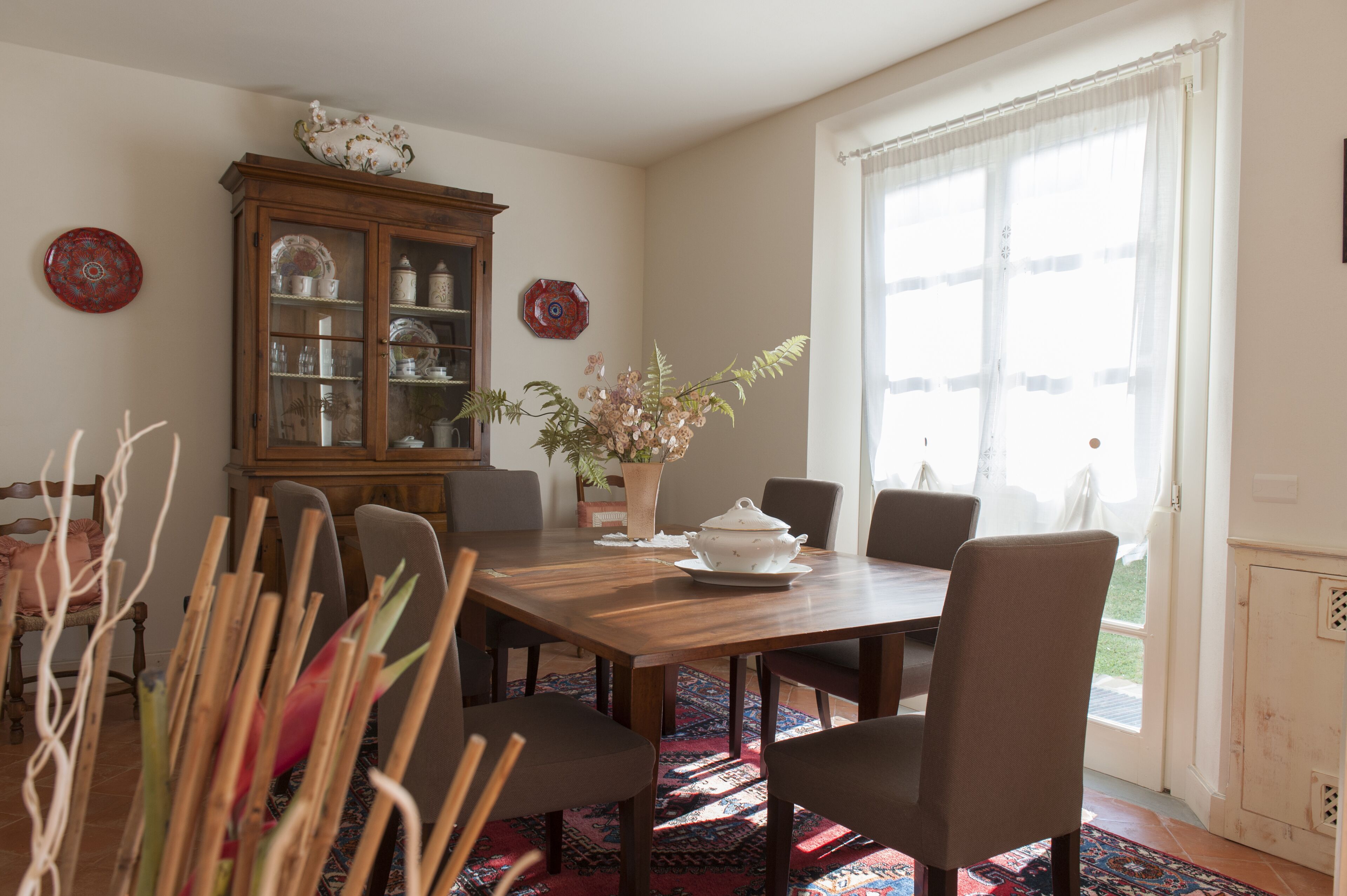 A dining area with a wooden table, six chairs, a cabinet with glass doors, a potted plant, and a large window with sheer curtains.