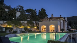A swimming pool with a curved edge, a building with arched doorways, and a hillside with trees. - Villa Caiano (Londa)