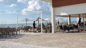 Ein Essbereich im Freien mit Tischen, Stühlen und Sonnenschirmen, mit Blick auf Strand und Meer.