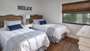 Ein Schlafzimmer mit zwei Betten, einem Nachttisch mit Lampe, einem Schild mit der Aufschrift 'RELAX' und einem Fenster mit Bambusrollo.