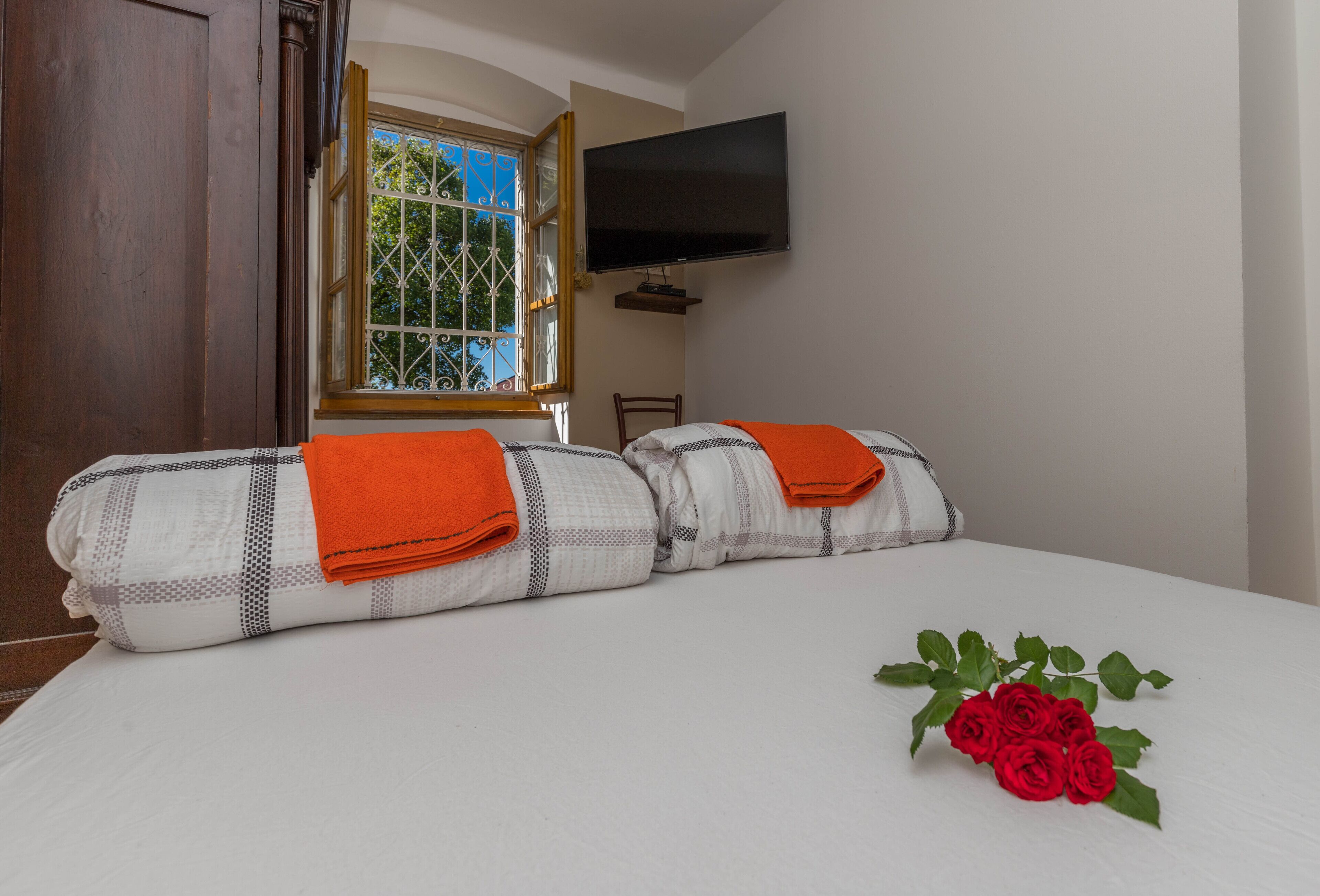 A neatly made bed with a checkered blanket and orange towels, a bouquet of red roses, a television mounted on the wall, and a window with a metal grille.