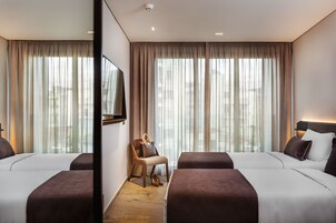 A hotel room with two beds, a chair, a television, and large windows with curtains.