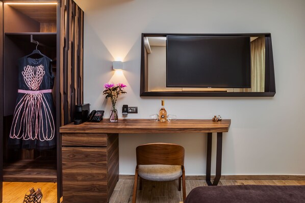 A hotel room with a wooden desk, a chair, a TV mounted on the wall, a dress on a hanger, a vase with flowers, and a telephone.