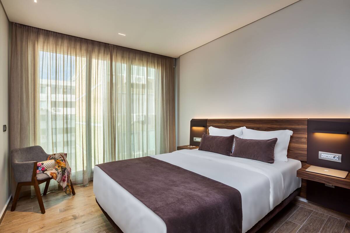 Une chambre d’hôtel moderne avec un grand lit, une table de chevet avec une lampe, une chaise recouverte d’un plaid à motifs floraux et une vue dégagée à travers des rideaux légers.