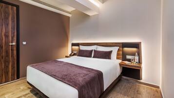 A hotel room with a large bed, a wooden headboard, a bedside table with a lamp, and a nightstand with a phone and a bottle.