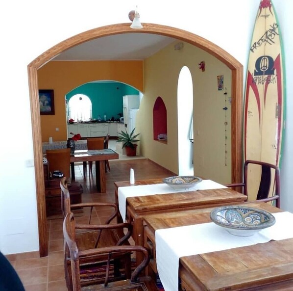 A dining area with wooden tables and chairs, a potted plant, and a surfboard on the wall.