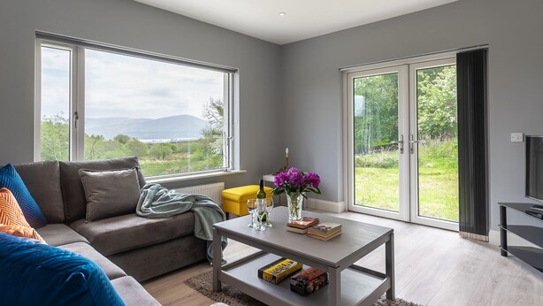 Ein modernes Wohnzimmer mit Sofa, Couchtisch und Blick auf Grünflächen und Berge durch große Fenster.