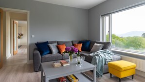 Ein modernes Wohnzimmer mit einem grauen Sofa, farbenfrohen Kissen, einem Couchtisch mit Büchern und Gläsern sowie Blick ins Grüne durch ein großes Fenster.