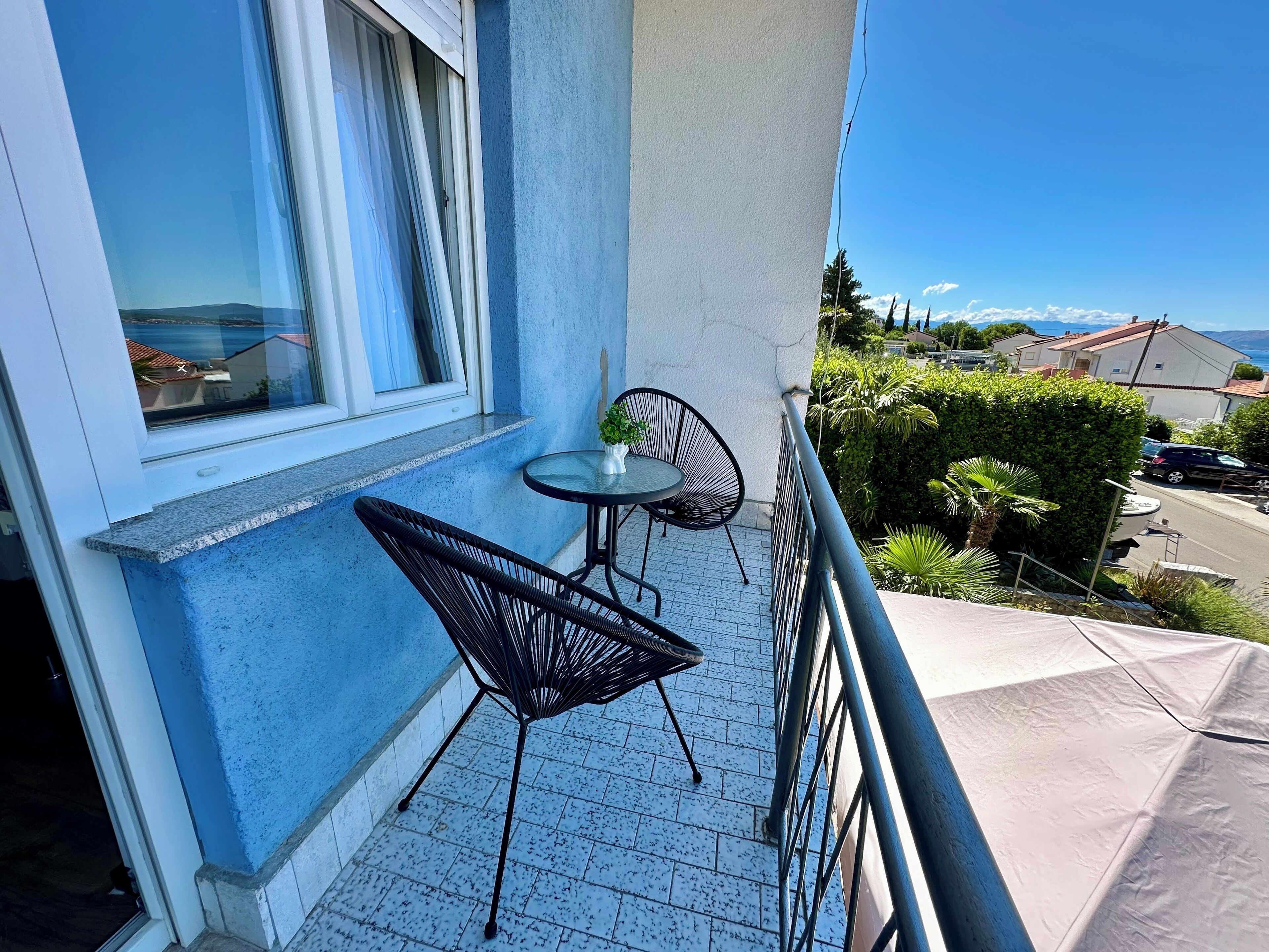 Uma varanda com uma mesa pequena e duas cadeiras, uma janela com cortinas e vista para o mar e casas.